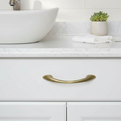 Close-up of a brushed antique brass cabinet handle on a white drawer with a marble countertop and sink in the background.