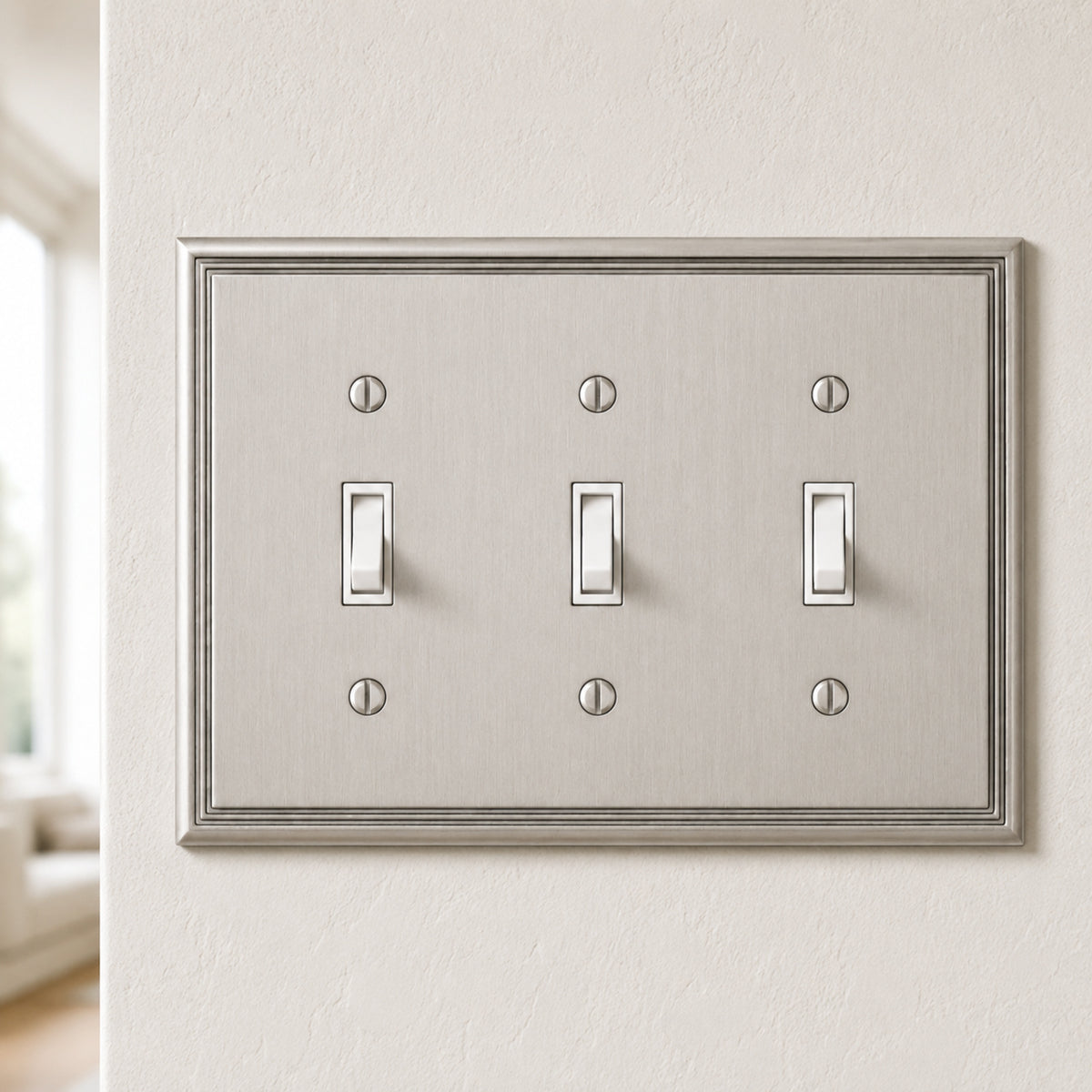 A three-light switch plate cover with a brushed silver metal finish and white toggle switches, mounted on a textured beige wall.
