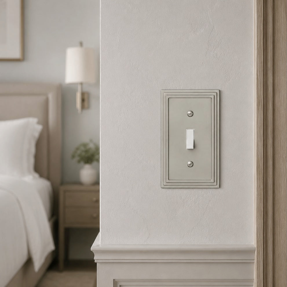 A single light switch plate with a vertical toggle, shown mounted on a textured off-white wall in a softly lit bedroom setting.