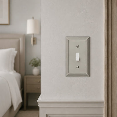 A single light switch plate with a vertical toggle, shown mounted on a textured off-white wall in a softly lit bedroom setting.