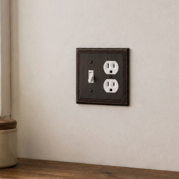 An oil rubbed bronze decorative electrical outlet cover plate with a single toggle switch and dual outlet, mounted on a textured beige wall above a rustic wooden counter.