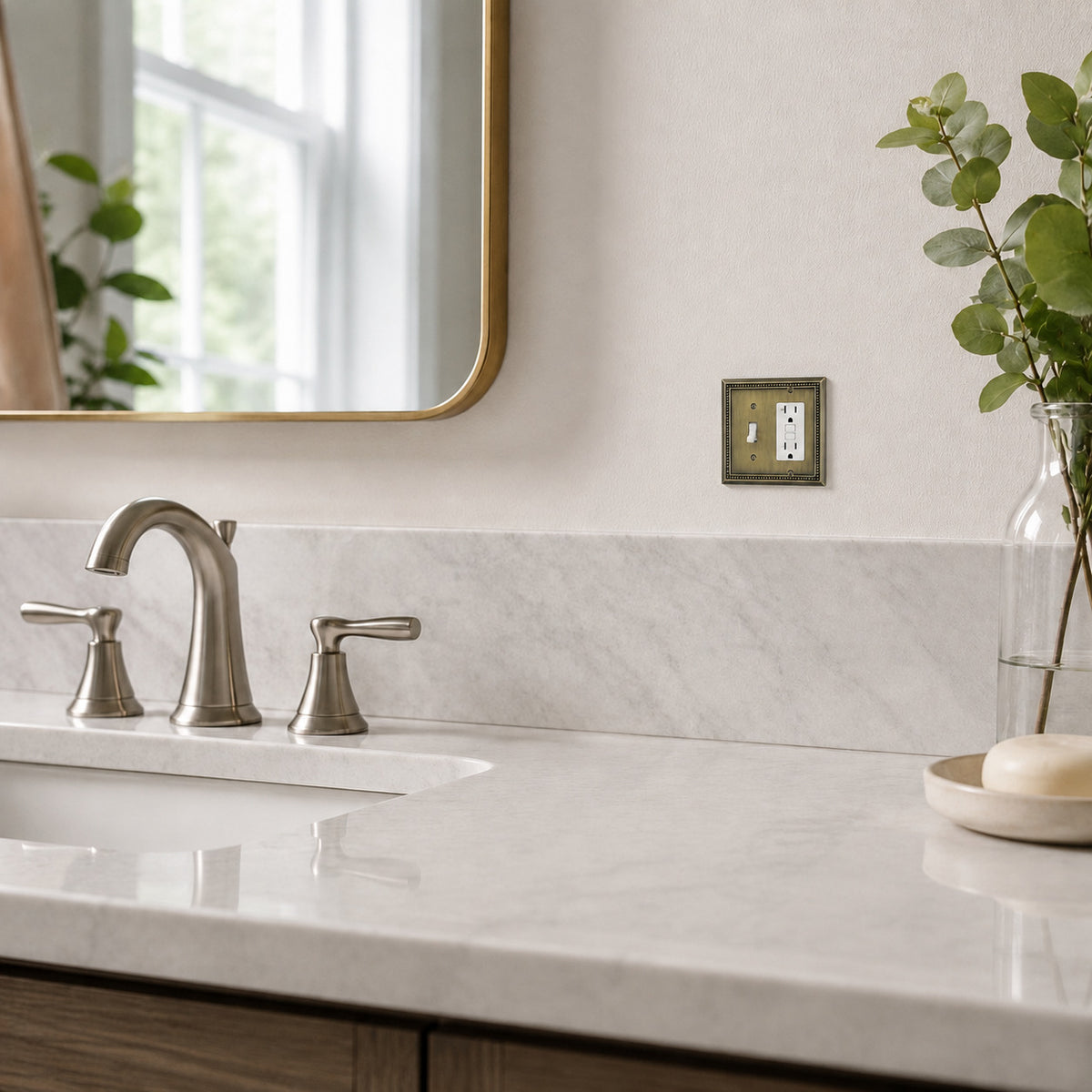 A bathroom vanity scene with a white marble countertop, brushed nickel faucet, wall mirror, and a small square wall-mounted fixture on the beige wall. A glass vase with greenery and a bar of soap sit on the counter.